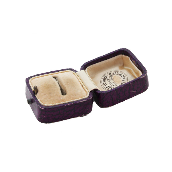 Open purple jewelry box with beige interior, containing a ring, on a white background.